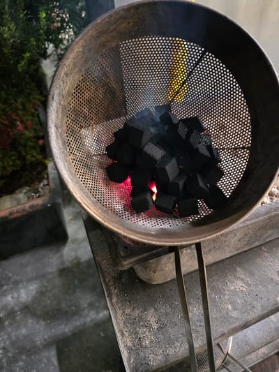 A close-up, high-angle shot captures a metal perforated basket filled with black, cube-shaped charcoal pieces. The charcoal is beginning to glow red hot from underneath, indicating it is being heated. The basket is resting on a dark metal stand, and a pair of metal tongs are visible on the right side of the stand. The background is out of focus, showing some greenery, likely plants in planters, and a dark, wet-looking ground surface. The scene appears to be outdoors, possibly a patio or balcony area, in Burbank, United States of America. There are no people visible in the image.