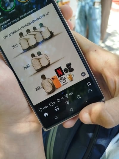 The image shows a hand holding a smartphone, displaying a social media post. The post features a drawing that illustrates a progression of friendships over time, starting with a group of four people in 2015, then two people in 2020, and finally one person with a phone in 2026, suggesting a decline in social interaction. Various icons are also visible on the screen, including Netflix, ChatGPT, dumbbells, and a coffee cup. The bottom of the screen shows social media engagement metrics like likes and comments. The background of the image is blurred but appears to be an outdoor setting with a person in light blue jeans in the distance. There is no indication of the image being taken in Goght, Armenia.