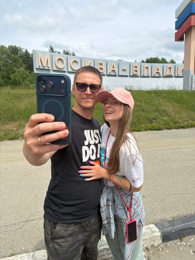 In a medium shot, a young couple takes a selfie in front of a sign that reads "MOSKVA-VLAD." The man, on the left, wears sunglasses and a black t-shirt with "JUST DO IT" printed on it, and camouflage pants. He is holding a dark blue smartphone, taking the photo. His left hand, which wears a gold wedding band, rests on the woman's shoulder. The woman, on the right, smiles as she leans into the man. She has a pink baseball cap on her head, and wears a white t-shirt with a graphic design, and a light blue plaid shirt tied around her waist. Her right hand, painted with bright pink and blue nails, is placed on the man's chest. A red cord hangs from her neck, attached to a pink phone case. The couple stands on a paved area next to a grassy slope. The sign is large and illuminated, with the letters outlined in gold. A tall, colorful monument stands to the right of the sign, partially visible in the frame. The sky is overcast. The location appears to be a roadside in Nikolaevka, Russia, indicated by the sign.