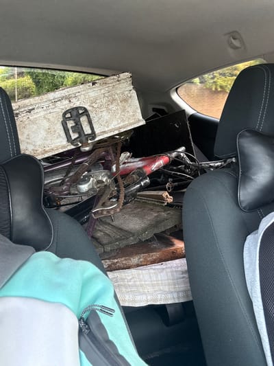 This vertical, eye-level image captures the cluttered interior of a vehicle, likely a car, from the perspective of a front passenger or driver. The main subjects are various objects being transported, including at least two bicycles and several pieces of wooden planks or crates, packed into what appears to be the back seat area.

One bicycle is clearly visible towards the top center, featuring a red and silver frame with the partial text "MOBI..." printed on its downtube. Another bicycle, predominantly purple or a dark color, is seen closer to the bottom center, showing a rusty chain and weathered pedals. One such pedal is prominently displayed on a dirty, white wooden plank or crate positioned in the foreground to the left. The other wooden pieces are rough, some appearing as old planks or fencing.

The car's interior features grey ceiling lining on the left, and dark grey fabric car seats with black leather headrests. White stitching is visible on the headrest and seat cushions. In the bottom right, the lap and lower body of a person are visible, wearing a soft garment in a light blue/mint green and white color block design, with a black zipper and patterned metal pull tabs.

Through the car window on the left, bright natural light illuminates lush green foliage and what appears to be a fence or building in the background, indicating it is daytime. The overall scene suggests the activity of transporting multiple items, including old bicycles and wood, possibly for disposal, repair, or a project. Although the image itself provides limited location cues, the context places the scene in Boston, United Kingdom. No specific emotions are discernible from the image.