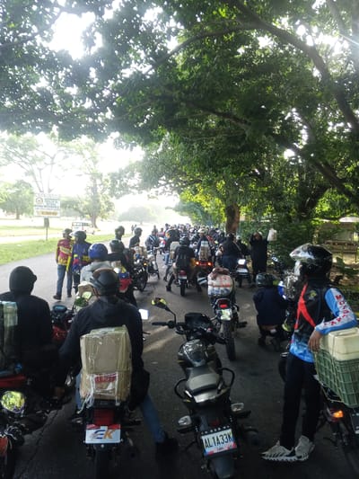 A group of motorcyclists are gathered on a paved road, likely in Campo Carabobo, Venezuela, as suggested by the presence of a sign in the background that reads "MILICIA BOLIVARIANA AGRUPAMIENTO POPULAR DE SEGURIDAD INTEGRAL INDEPENDENCIA ADI-454 LIBERTADOR A 150 MTS." The road is lined with trees, casting shadows and dappled sunlight onto the scene. The motorcyclists are wearing helmets and casual clothing, and many of their motorcycles are equipped with carriers, some laden with boxes. Some individuals are standing next to their bikes, while others are seated. The atmosphere appears to be one of a gathering or a planned event, with a substantial number of motorcycles parked and people congregating. The lighting suggests it is daytime, possibly morning or late afternoon, given the low angle of the sun filtering through the trees.