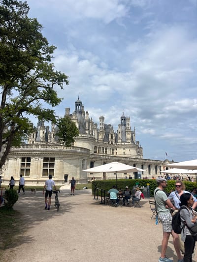The image captures the Château de Chambord in Chambord, France, on a partly cloudy day. The impressive architecture of the château, with its many towers and ornate stonework, is a prominent feature. People are present in the foreground and along the side, some walking and others seated at tables under umbrellas. A cyclist is seen with a bicycle. A large tree partially obscures the view of the château.