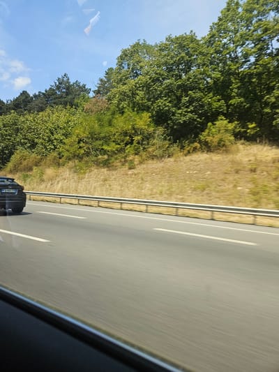 The image shows a road from the perspective of a vehicle driving on it. The road is paved and has white lane markings. To the left of the road, there are trees and a grassy area. The sky is blue with some clouds. The sun appears to be shining, suggesting it is daytime and the weather is clear.
A dark-colored car is visible on the road ahead, also moving in the same direction. The license plate of this car is partially visible and reads "FG 521 TN". The image is taken at an angle, possibly due to the movement of the vehicle it was captured from, or the camera itself.
The overall scene depicts a typical highway or main road environment, likely in a rural or suburban setting given the presence of trees and vegetation. The motion blur suggests the vehicle is in transit at a moderate to high speed. There is no clear indication of people in the image, only the car. The location context provided is Châtenay-Malabry, France.
