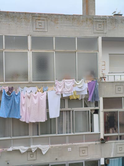 A medium shot captures a concrete building facade in Nazaré, Portugal, with laundry hanging on a clothesline in front of windows. Two seagulls are perched on the rooftop in the upper right corner of the frame. The building exhibits architectural details such as recessed square motifs on its concrete exterior. The windows appear to be double-paned and offer obscured views of the interior. The laundry includes various items of clothing in different colors and patterns, including a blue gingham sheet, pink patterned fabrics, white towels, and a purple garment. The clothesline is supported by clothes pegs. The sky is overcast.