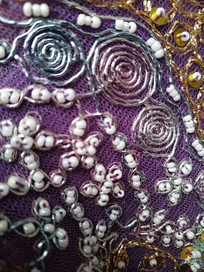 This close-up image captures an intricate detail of a richly embroidered textile, likely a traditional garment or decorative piece, from Houaykhang, Laos. The dominant element is a vibrant, deep purple fabric, which serves as the background for elaborate embellishments.
The textile is adorned with a variety of metallic threads, beads, and sequins. Silver-colored metallic thread forms swirling, spiral patterns, resembling coiled springs or abstract floral motifs, along with more fluid, curved lines. These silver patterns are often highlighted by clusters of small, opaque white beads, many of which appear to have a tiny purple stitch or center, suggesting they are secured with a contrasting thread. These beads frequently form small, multi-lobed floral or leaf-like shapes.
Towards the right side of the frame, gold-colored metallic thread embroidery is visible, creating a distinct, somewhat geometric pattern that interweaves around small, circular, gold-colored sequins. These sequins are typically secured at their centers with a stitch, and the gold threadwork often outlines them. Additional white beads are interspersed throughout both the silver and gold sections of the embroidery, arranged in linear sequences or small groups.
The image emphasizes the texture and shimmering quality of the metallic threads and sequins against the woven purple fabric, showcasing detailed and meticulous handcraftsmanship. No people, activities, or text are visible in this static, macro shot.