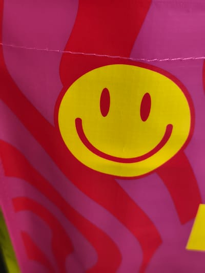 This is a close-up, indoor, still image of a pink and red striped fabric with a large yellow smiley face graphic. The smiley face is in the center of the frame, with two red oval eyes and a red curved smile. The fabric has a grid-like texture. There is a visible seam across the top of the image, which appears to be a stitched hem or joining of fabric pieces. The image appears to be of a bag or other fabric item, as suggested by the texture and the seam. The lighting is bright and even, suggesting it was taken in a well-lit indoor environment. There is no discernible time of day, weather, or location cues beyond the provided context of Troisdorf, Germany. The overall emotion conveyed by the smiley face graphic is positive and cheerful. No text is visible in the image.