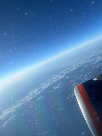 The image presents an aerial view captured from an aircraft, looking downward and towards the distant horizon over a vast landmass. This scene is situated over Russia, with the provided context placing the location above Berezovaya Roshcha. The upper two-thirds of the frame show a clear, deep blue sky, which gradually lightens to a pale blue-white at the horizon line. Scattered across the sky and upper landscape are numerous small, out-of-focus circular white specks, likely particles on the aircraft window. Below the horizon, the terrain consists of extensive dark-colored areas, presumably forests, interspersed with irregular and geometrically shaped lighter patches consistent with snow-covered ground or agricultural clearings. In the lower right corner, a section of an aircraft engine nacelle is visible. It is predominantly dark blue or black, featuring a prominent horizontal orange-red stripe and a metallic silver leading edge. A small white projection, possibly a sensor or probe, extends from the upper surface of the nacelle. No people, specific actions, or direct interactions are visible within the frame.