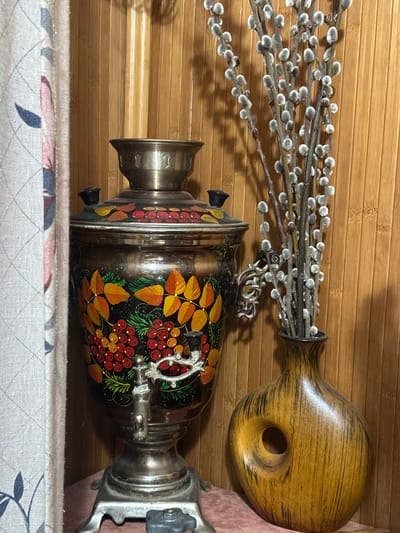 A samovar with floral painted decoration and a faucet stands to the left of a wooden vase with a large circular opening, holding a bouquet of pussy willow branches. To the far left, a curtain with a floral pattern is partially visible. The background is a wooden wall with vertical planks. The scene is indoors, likely in Chornomors'ke, Ukraine, given the contextual information.