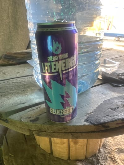 A tall, purple and blue energy drink can, branded "LIT ENERGY" and "BLUEBERRY," sits on a rough, weathered wooden surface. Behind the can is a large, clear plastic bottle with a ribbed texture, likely containing water. The background is slightly out of focus but suggests an outdoor, possibly industrial or rural setting, with hints of sunlight and a sandy or dusty ground cover. The scene appears to be in or around the village of Chikhchino, Russia.