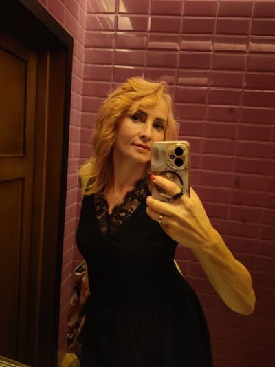 A middle-aged Caucasian woman with blonde hair styled in loose waves is taking a selfie in what appears to be a restroom. She is wearing a black dress with a lace neckline. She is holding a smartphone in her right hand, which is also adorned with rings. The background consists of pink tiled walls and a wooden door. The image does not provide any information that would allow for the determination of the city of Chelganovo, Russia, or any specific location.