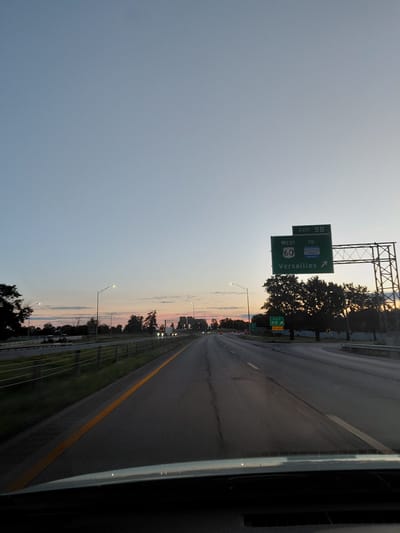 The image depicts a highway scene at dusk in Lexington, United States of America. A highway sign indicates "EXIT 5B WEST TO 60 Versailles" with an arrow pointing to the right. Another smaller sign below shows "30 MPH". The highway is paved with lane markings and a yellow line. Streetlights illuminate the scene, and trees line the side of the road. In the distance, vehicles can be seen on the highway. The sky is a gradient of light blue to pink, indicating sunset or sunrise. The perspective is from inside a vehicle, looking forward.