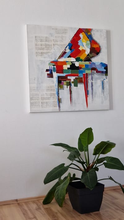 The image shows a framed abstract painting hanging on a white wall. The painting is predominantly white with colorful abstract shapes and lines in red, yellow, blue, green, and black. Part of the painting appears to be collaged with what looks like text or newspaper clippings. To the right of the painting, a dark planter holds a lush green plant with large, elongated leaves. The plant is positioned high on the wall and cascades downwards. The background wall is white, and to the far right, a section of a light brown wooden floor or paneling is visible, suggesting an indoor setting. The lighting appears to be neutral, and there are no explicit indicators of time of day or weather. The overall scene is a still life composition of art and nature within a domestic or gallery space. There is no discernible text that can be clearly read within the image. The location context provided is Ichenhausen, Germany, but there are no visual cues in the image to confirm this specific location.