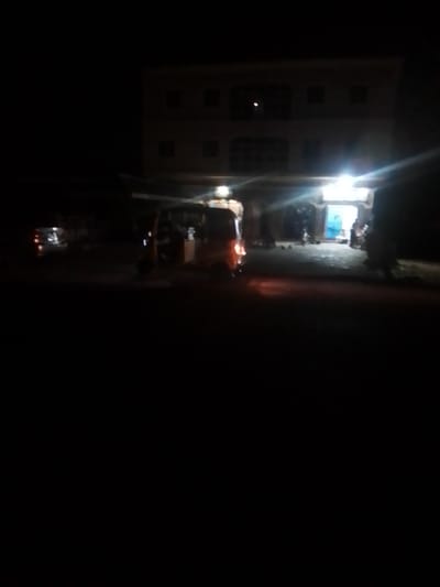 The image depicts a nighttime scene on BYE PASS, Nigeria. In the foreground, a three-wheeled vehicle, possibly a tuk-tuk, is visible, illuminated by a bright overhead light source. To the left, a car with its taillights on is parked. In the background, a multi-story building is partially lit, with lights visible in some of its windows. The overall scene suggests a commercial or residential area that is active at night. The environment appears to be a street or road in front of a building, with some debris or vegetation on the ground. The lighting is predominantly artificial, coming from the building and streetlights, creating a somewhat hazy and atmospheric effect.