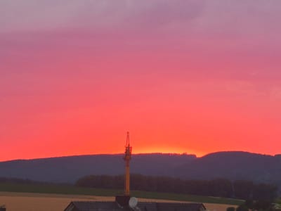 A dramatic sunset paints the sky in vibrant hues of pink and orange over a rural landscape near Lügde, Germany. Silhouetted against the colorful sky are rolling hills covered with trees, with fields and rooftops in the foreground. A tall yellow construction crane stands prominently in the mid-ground, its structure stark against the glowing sky. The overall scene is one of natural beauty and industrial presence coexisting under an extraordinary sky.
