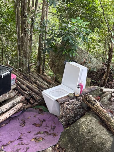 The image depicts a disorganized, makeshift outdoor area in a wooded environment. Location: Moo 7, Thailand. Dominating the scene is a structure constructed of logs, supporting various items. A white plastic box, partially open, rests atop this platform. Next to it are cleaning supplies, a small container with pebbles and a potted plant. Beneath the structure is a black plastic container. A purple mat is positioned to the right of the wooden structure. The surrounding environment is characterized by abundant foliage and large rocks.