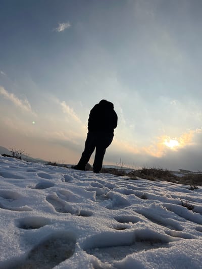 A lone individual is depicted standing on a snow-covered uphill slope in Talfita, Syria. The person, viewed from behind and angled slightly right, is silhouetted against a bright sky, wearing dark, heavy winter clothing, including a thick jacket and trousers. The foreground consists of granular snow marked with numerous distinct boot prints, with patches of dry vegetation and exposed earth visible throughout. The sky transitions from a pale blue at the top to a warm, yellowish-orange hue near the horizon. The sun, partially obscured by clouds, creates a bright glare on the right side of the frame. Scattered wispy clouds are present, and a green lens flare is visible in the upper left quadrant of the sky. In the distant midground, faint outlines of structures or utility lines are discernible on the left, and a building-like structure appears on the far right.