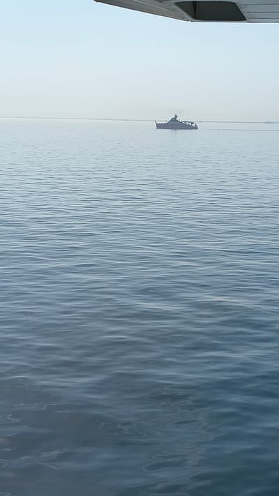 A medium-shot, eye-level, horizontal, slightly high-angle, natural light, outdoor, daytime photograph shows a large, dark-colored ship sailing on calm, blue water. The ship is located in the middle ground of the image, positioned slightly to the right of the center. It has a dark superstructure and appears to be a commercial or military vessel. The horizon line is visible in the far distance, under a pale, hazy sky. The foreground and middle ground are dominated by the surface of the water, which exhibits gentle ripples and reflections. A portion of a light-colored wing of an aircraft is visible in the upper left corner of the frame, suggesting the photograph was taken from an airplane. The scene is devoid of any human figures, and the environment is entirely natural, with no man-made structures or urban elements visible, except for the ship and the wing of the aircraft.