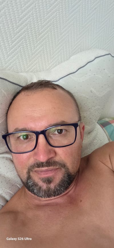 A man with short, receding brown hair and a goatee is lying in bed, looking at the camera. He is wearing blue-rimmed glasses. The bed appears to be in a bedroom in a home. The background is a white textured surface, possibly a headboard or wall. A portion of a pillow with a blue and white checkered pattern is visible on the right side of the image. The lighting suggests a natural light source, possibly from a window. The text "Galaxy S26 Ultra" is visible in the bottom left corner of the image. No specific location in the city of Evergem, Belgium is depicted.
