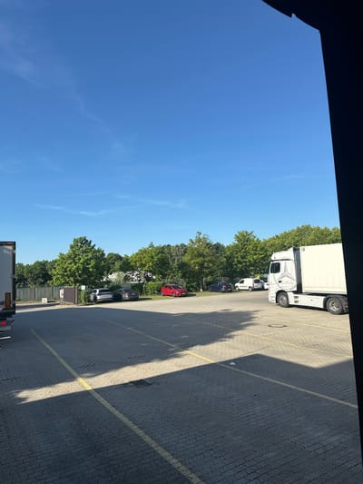 A bright, clear blue sky fills the upper portion of the image. Below, a paved parking lot is visible, marked with yellow lines. In the distance, a row of lush green trees forms the backdrop. Parked cars are scattered across the lot, including a red car, a dark blue car, a silver SUV, and a gray sedan. A white van and a large white semi-truck with a trailer are also present. The semi-truck has a Mercedes-Benz logo on its cab. The ground is made of interlocking pavers, and shadows are cast across the surface. The image appears to be taken from an elevated, shaded vantage point, possibly an open doorway or window. The scene depicts an outdoor parking area, likely at a business or industrial site.