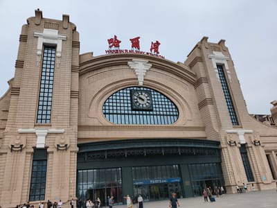 The image shows the Harbin Railway Station in Harbin, China, on a cloudy day. The station is a large, ornate building with a beige facade and prominent architectural features, including tall, rectangular windows with dark panes and decorative white elements. Above the main entrance, a large, circular clock is visible within an arched window. Red Chinese characters, which translate to "Harbin Railway Station," are displayed prominently on the building's roof, along with the English text "HARBIN RAILWAY STATION."
In front of the station, a paved area is occupied by a moderate number of pedestrians. Some are walking, while others appear to be waiting or observing. Several individuals are seen with luggage, suggesting they are travelers. The people are dressed in casual attire, typical of a warm day. The scene is dynamic, with people moving in various directions, creating a sense of activity and transit. The overall impression is that of a busy public transportation hub.