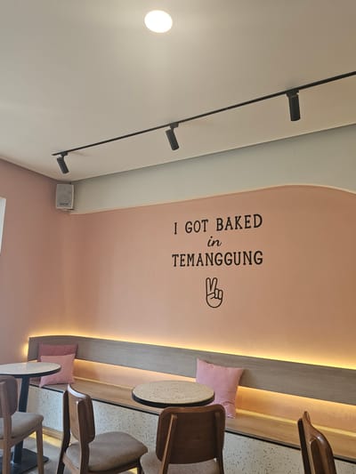 The image depicts the interior of a cafe in Banyuurip, Indonesia. The wall is painted a soft pink and features black lettering that reads "I GOT BAKED in TEMANGGUNG" with a peace sign graphic below it. A long, light-colored wooden bench with a speckled front is built into the wall, illuminated from above by a warm LED strip light. Two pink cushions rest on the bench. In front of the bench, two round tables with speckled tops are visible, each accompanied by wooden chairs with grey upholstered seats. One table is closer to the left edge of the frame, and the other is slightly to the right of center. The ceiling is white with a track lighting system featuring three black spotlights. A white speaker is mounted on the wall in the upper left corner. The overall atmosphere appears to be modern and casual, typical of a cafe setting.