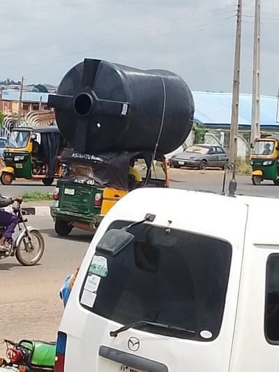 A large black water tank is being transported on top of a yellow and green tuk-tuk in Oja Oba, Oko Erin, Nigeria. The tuk-tuk has a black tarpaulin covering its interior, over which the tank is secured. Text is visible on the tarpaulin, including "ASIWATU B" and "NICE (4)". A white van is partially visible in the foreground, obscuring the back of the tuk-tuk. Another tuk-tuk, yellow and green, is visible to the left of the tank-laden vehicle. In the background, a grey sedan is parked on the side of the road, and a person is riding a white motorcycle with a red taillight. Electrical poles and overhead wires are also present in the background. The sky is overcast.
