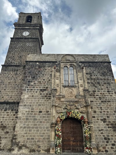 This is a low-angle, full-shot, exterior, eye-level photograph of the Parish of San Andrés Apóstol, located in San Andrés Calpan, Puebla, Mexico. The church features a large, weathered stone facade with a prominent bell tower on the left. The tower has a clock face visible, and a bell within an open belfry. The main entrance is a large, arched wooden door adorned with an elaborate floral arrangement of white and red flowers. Flanking the door are carved stone pillars and decorative stonework above the arch. A large window with mullions is positioned above the doorway. The sky above is cloudy and overcast. The image depicts the church from a frontal perspective, emphasizing its architectural details and the festive floral decorations.