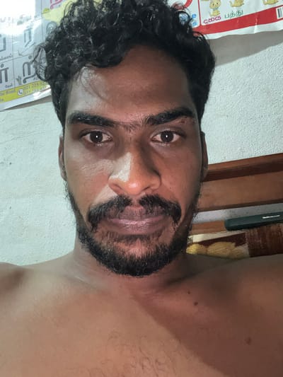 The media file presents a portrait of a man, likely taken in Ulla, Sri Lanka, presented in a rotated orientation (90 degrees counter-clockwise). The man, with a dark complexion, curly black hair, and a thick beard, is looking directly at the camera with a neutral expression. A distinct white patch is visible on his nose, suggesting the application of sunblock or a similar cosmetic. He appears to be shirtless, with his bare chest and shoulder visible.

The background indicates an indoor setting. To the left (when viewed in correct orientation), a textured white wall is visible. To the right, there is a wooden frame or panel, and a section of patterned fabric, possibly a rug or cushion. Behind the man's head, part of a calendar or poster is visible, featuring cartoon cats and text primarily in Tamil. Visible Tamil phrases include "பூனை பந்து" (Poonai Pandhu), which translates to "Cat ball." Partial phone numbers such as "021 225 55" and "077 284 63" are also present. Other discernible Tamil words include "அங்காடி" (Angadi - market/shop) and "காலை" (Kaalai - morning/bull). The lighting appears to be from an indoor source.