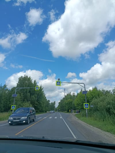 The image depicts a road in the city of Тополиный, Russia, under a bright blue sky with scattered white clouds. The road is paved and has two lanes of traffic. Pedestrian crossing signs, recognizable by their yellow triangular shape with a blue pedestrian silhouette, are visible on both sides of the road, mounted on poles. Some of these signs appear to be solar-powered, indicated by the solar panels attached to the poles.
A dark-colored minivan is driving in the left lane, heading towards the viewer. The minivan's headlights are illuminated. Several other vehicles are visible in the distance on the right lane, also moving away from the viewer. The road is bordered by lush green trees and vegetation on both sides. On the right side of the road, near a pedestrian crossing, a small group of people is standing, some appearing to be waiting. The foreground of the image is dominated by the dashboard and windshield of a vehicle, suggesting the image was taken from inside a car. The overall scene is that of a typical suburban or rural road on a sunny day.