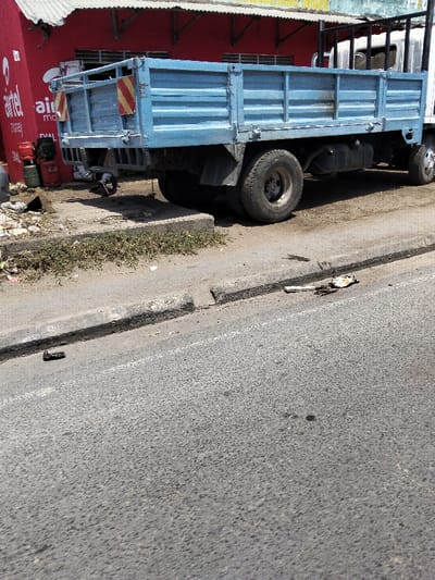In Mkomani ward, Kenya, a light blue flatbed utility truck is parked on a dirt verge alongside an asphalt road, positioned with its rear facing the viewer and its front towards the left of the frame. The truck features a weathered metal flatbed with horizontal slats, dual rear wheels, and two rectangular yellow and red diagonally striped reflective markers on the rear structure.
Behind the truck, a red building with a white corrugated metal roof is visible. A red "Airtel money" sign is partially displayed on the building's corner. Several red and green gas cylinders are placed on the ground next to the building. The dirt verge exhibits sparse green vegetation and accumulated debris.
A concrete curb separates the verge from the asphalt road. A second, lower concrete curb runs parallel to the main curb further onto the road surface. The asphalt road shows minor wear, scattered dark stains, and debris. A dark, irregular object is situated on the road in the foreground, while a flattened white object with dark residue lies near the lower curb. No individuals are present, and no actions or interactions are depicted.
