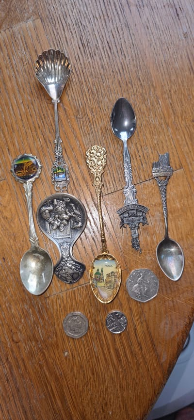 The image shows a collection of souvenir spoons and coins arranged on a wooden surface. The spoons are of various designs, some featuring enamel artwork of cityscapes or buildings, while others have ornate metalwork. One spoon has a shell-shaped bowl, and another appears to depict a scene with figures. The coins are scattered among the spoons, with visible designs and denominations. The wooden surface has a visible grain pattern. The image does not contain any recognizable features that would indicate a specific location in the city of Marum, Netherlands.