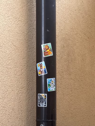 A black, cylindrical pipe stands vertically against a textured, tan-colored wall. Four stickers are affixed to the pipe, arranged from top to bottom. The topmost sticker features a cartoonish lion with a yellow mane and chest, depicted with its mouth open. Below that is a sticker of a cartoon reindeer with antlers and a brown torso, holding yellow orbs. The third sticker shows a dalmatian dog wearing a blue collar. The bottom sticker depicts a dark, hooded figure with a white outline. The scene appears to be outdoors, possibly in a public area within the city of Suceava, Romania, given the context provided.