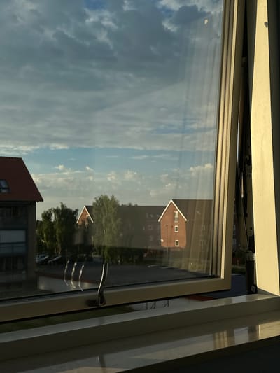 A view through an open window in Roskilde, Denmark, reveals a residential area bathed in the warm light of late afternoon sun. The window frame, a pale beige color, is open at an angle, allowing a glimpse of the exterior. The lower part of the windowpane shows reflections of buildings and trees, suggesting an urban or suburban setting.
Beyond the window, a two-story building with a red-tiled roof and grey facade is visible on the left. To the right of this, several trees with lush green foliage stand tall. Further in the background, another building with a red brick facade and a prominent gable roof is seen, its windows reflecting the sky. The sky itself is a mix of blue and grey clouds, with some patches of brighter sky suggesting intermittent sunlight.
The interior windowsill in the foreground is a pale color, reflecting the light and appearing glossy. A metallic window latch is visible on the lower part of the window frame. The overall impression is one of a calm, everyday scene, capturing a moment of natural light illuminating a residential landscape.