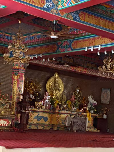 The image shows the interior of a Buddhist temple, possibly located in Việt Trì, Vietnam, given the context. The ceiling is elaborately decorated with red beams and intricate colorful murals featuring swirling patterns, floral motifs, and what appear to be Tibetan or Sanskrit script in yellow. Two ornate golden chandeliers hang from the ceiling, and a ceiling fan is visible.
The walls are constructed of brick. In the center of the scene is a raised altar adorned with various religious statues and artifacts. A prominent golden statue of a multi-armed deity is central, surrounded by smaller golden figures and decorative boxes. To the right of this, a white statue of a seated figure with many arms is visible, along with floral arrangements. In front of the altar is a red carpet that covers the floor. To the left of the altar, a large, decorated pillar with red and gold motifs supports the ceiling structure.
Further to the left, another altar or shrine structure is visible, featuring a painting of an elephant. The overall atmosphere is one of spiritual reverence and artistic richness. No people are visible in the image.