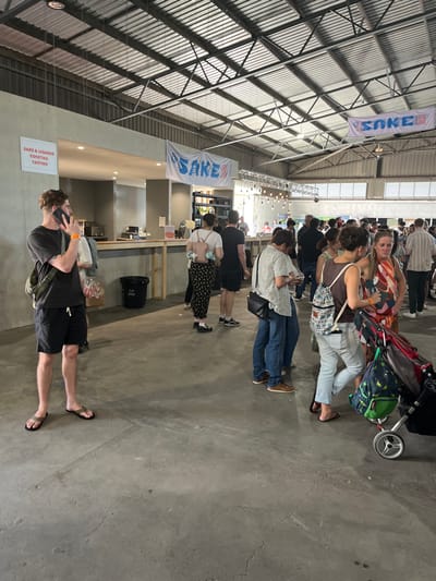 The image shows a large, open indoor space with a concrete floor. The space appears to be part of a large warehouse or industrial building, with a metal truss roof.  The space is populated with people, mostly adults, who are standing and talking. There is a bar-style counter to the left of the image.  The counter has signage for sake and liquor tastings. A group of people are gathered around a small bar counter, while others are standing in groups or walking around. Several people are holding drinks and some are wearing blue wristbands. There is a sign above the bar counter that says "SAKE." A sign near the roof says "SAKE." There is a black bin next to the counter. In the foreground, one person is on the phone. There are two baby strollers in the image, both of which have backpacks attached to them. The image was taken at a sake bar at the "SAKE" festival at 400 Montague Rd, West End, Hamilton, Queensland 4101, Australia.  (latitude: -27.439, longitude: 153.075).