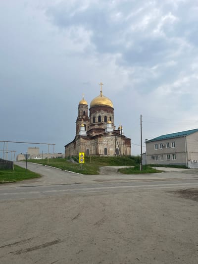The image depicts a large, old, brick church with golden domes in Maminskoe, Russia. The church is situated on a grassy hill. To the right of the church is a modern, light-colored brick building with a green roof. In the foreground, a dirt road curves around the base of the hill, with tire tracks visible on the surface. A yellow traffic sign with a pedestrian symbol and a "40" speed limit is posted near the road. The sky is overcast with gray clouds. There are no people visible in the scene.