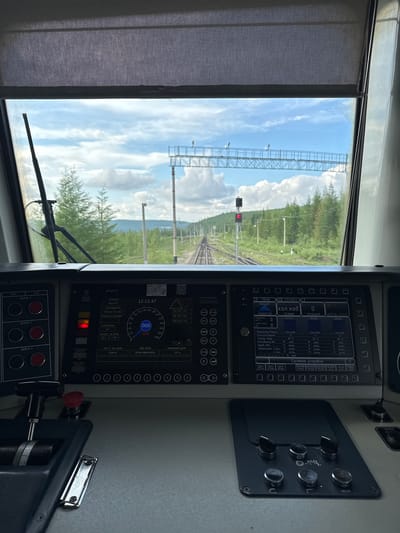 The image depicts the interior of a train cab, looking out through the windshield at a railway track stretching into the distance, surrounded by a forest under a cloudy sky. The foreground is dominated by the train's control panel, featuring various buttons, levers, and digital displays. On the left side of the control panel, there are several large circular buttons labeled "ПУСК" (Start), "СТОП" (Stop), and "ДИЗЕЛЬ 1", "ДИЗЕЛЬ 2", "ДИЗЕЛЬ 3". Below these are two circular buttons and a lever. A red emergency stop button is also visible. The central part of the control panel displays a digital screen showing train information. Key readings include "СТАНЦИЯ" (Station) as "БЕЗ СНС" (Without SNS), "КООРДИНАТА" (Coordinate) as "0341км 7лк 50м" (0341km 7th side 50m), "ВРЕМЯ ПО ГРАФИКУ" (Time according to schedule) as "0.0", and "УСКОРЕНИЕ" (Acceleration) as "0.0". It also shows "КАНАЛ № ПУТИ" (Track channel) as "25 Гц" (25 Hz) and "1 НП" (1 NP). A speed indicator dial shows a reading around 20 km/h, with the number "020" highlighted in a circle. Below this, "ФАКТИЧЕСКАЯ СКОРОСТЬ" (Actual speed) displays "0.00 km/h". Further displays read "РАССТ. ДО ЦЕЛИ" (Distance to destination) as "0184 M" (0184 meters), "ВИД ЦЕЛИ" (Type of destination) as "РЕЛЬСОВАЯ ЦЕПЬ" (Track circuit), and "НАЗВАНИЕ ЦЕЛИ" (Name of destination) as "25 Гц" (25 Hz). To the right, another digital display screen shows "ХОЛ. ХОД" (Idle mode) with multiple entries, including "Мощность/Режим" (Power/Mode) as "0", "Темпер. Воды" (Water temperature) as "81 °C", "Боксование/Бз" (Skidding/Bz) as "нет" (no), "Заторможен 1 мед" (Braked 1 second) as "2 мед" (2 seconds), and "Наклон/Бакет" (Incline/Bucket) as "5 Гр" (5 degrees). Other parameters like "Датчик" (Sensor), "ЭКСПЛУАТАЦИИ" (Operation), and "СИСТЕМА ИСПРАВНА" (System is OK) are also visible. The view through the windshield shows a double railway track disappearing into the horizon. The tracks are flanked by a dense forest of coniferous trees. In the distance, a signal light is visible, currently showing red. Overhead, a railway signal gantry with lights spans the tracks. The sky is partly cloudy with a bright, diffused light, suggesting daytime. The context provided places this scene in Zolotinka, Russia. The operational status and labeling of the control panel suggest this is a working train cab, likely in a remote or rural area given the surrounding forest. There are no people visible in the image. The overall impression is one of a functional and operational train environment, poised for