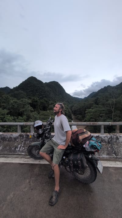A man with medium-length, light brown hair and a beard, wearing sunglasses on his head, a light grey t-shirt, and olive green shorts, sits on a dark green motorcycle on a bridge. The motorcycle is equipped with saddlebags and several bags strapped to the back. A helmet rests on the front of the motorcycle. The man is leaning forward with his right arm resting on the motorcycle's seat and his left hand on the handlebars. He is looking upwards and to the right. In the background, a river flows between lush green, jungle-covered mountains under a cloudy sky. The bridge railing is visible in the foreground. The location is identified as Thượng Trạch Commune, Vietnam.