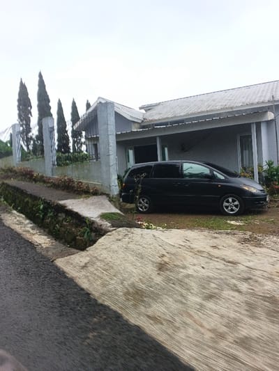 The scene depicts a static exterior view of a residential property in Masaiti, Zambia, under an overcast sky. In the foreground, a dark asphalt road transitions into a textured concrete driveway leading up to the property. A concrete drainage ditch runs along the left edge of the driveway. The property features a single-story house with light grey walls and a light-colored, possibly corrugated metal, sloped roof. A covered porch area extends from the front of the house. A low concrete wall topped with a dark metal fence marks the property boundary, with a concrete pillar visible at the driveway entrance. Several tall, slender conifer trees are planted behind the fence on the left side of the property. A black minivan, identified as a Toyota Previa/Estima model with silver multi-spoke alloy wheels, is parked on a dirt patch in front of the house, facing left. No individuals or active subjects are present in the image.