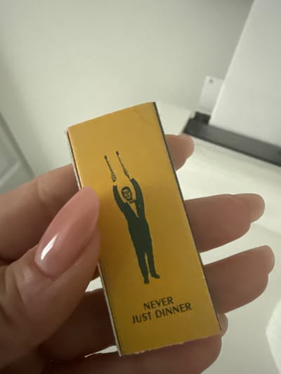 A close-up, slightly angled photograph captures a human hand holding a small, golden-yellow rectangular object, likely a matchbox or small container. The hand features light pink, almond-shaped, polished fingernails.
The held object is predominantly golden yellow with a distinct black graphic and text. The graphic is a silhouette of a person, possibly a man, standing with arms stretched upwards, holding two thin, stick-like objects. The figure's head displays some internal detailing. Vertically oriented on the right side of the object, the black text "NEVER JUST DINNER" is clearly visible.
The background is a bright, plain white or off-white surface, suggesting an indoor setting. A dark, linear element, possibly a shelf edge or a fixture, is partially visible in the upper left portion of the image. The lighting is bright and even, characteristic of an interior space. No specific activity beyond holding the object is depicted, and no emotions are discernible. The image provides no specific location cues for Davie, United States of America.