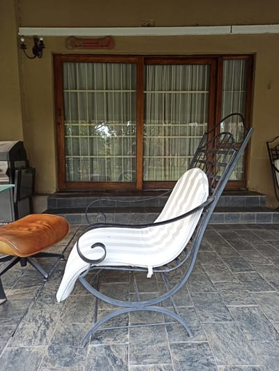 The image depicts an outdoor patio area with two pieces of furniture in the foreground. On the left is a brown leather ottoman with a black metal base. To its right is a black metal lounge chair with a white and grey striped cushion. The chair is angled, with its back facing away from the viewer. Behind the furniture, a wooden-framed sliding glass door with vertical metal bars on the exterior is visible. The door leads to an interior space with sheer curtains. The patio is paved with dark grey, textured tiles. In the background, a light-colored wall of a building is visible, with a decorative bone-shaped object mounted above the door and a dark metal sconce light fixture to the left. The scene appears to be in Randburg, South Africa, suggesting a residential or commercial outdoor living space.