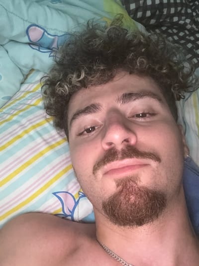 A person with curly dark hair and a beard lies on a bed, looking upwards towards the camera. The person is wearing a blue shirt and a silver chain. The bed is covered with a light blue sheet that has a pattern of colorful stripes and a drawing of a flower. The person's face is partially obscured by their hair. The lighting suggests it may be daytime, as there is no artificial light visible. The scene appears to be indoors, possibly in a bedroom. The person's expression is neutral.