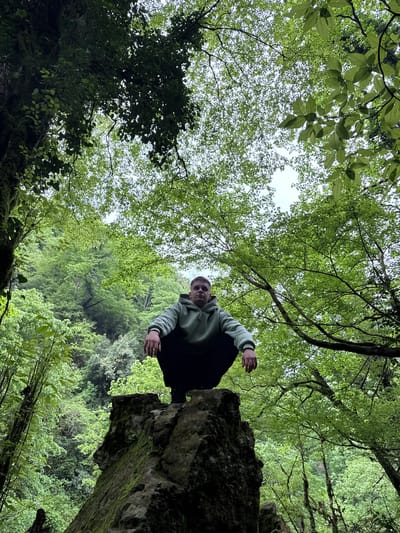 A low-angle shot depicts a person perched atop a moss-covered rock formation, with a dense forest canopy overhead. The person, wearing a light-colored hooded sweatshirt, dark pants, and a dark beanie, squats on the rock, looking directly down at the camera. The forest canopy fills most of the background, composed of green leaves. The location is inferred to be the city of Tsabal in Abkhazia.