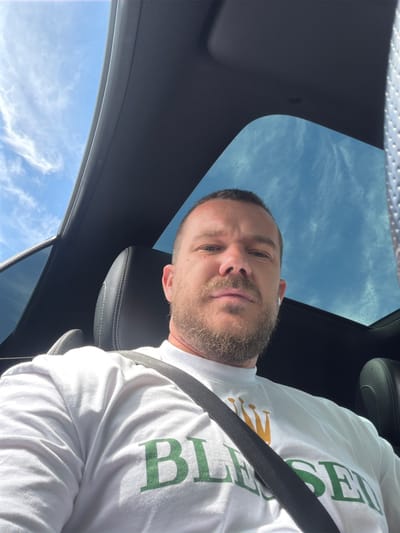 A low-angle, upward-facing selfie captures a man seated inside a vehicle, looking directly at the camera. The man, who appears to be in his 30s or 40s, has short hair, a light skin tone, and a neatly trimmed beard and mustache. His expression is neutral and direct.
He is wearing a white t-shirt with green text that prominently displays "BLESSED" and a golden crown-like graphic beneath the letter 'B'. A black seatbelt stretches diagonally across his chest and shoulder. The car's interior features dark-colored, possibly leather, seats with visible stitching and a dark grey or black roof lining.
Above the man, a sunroof offers a clear view of a bright blue sky with scattered white clouds, indicating it is daytime. Small water droplets are visible on the sunroof glass. A dark car pillar and a side window, possibly tinted, are also visible in the frame, confirming the setting inside a vehicle. The lighting is bright and natural.