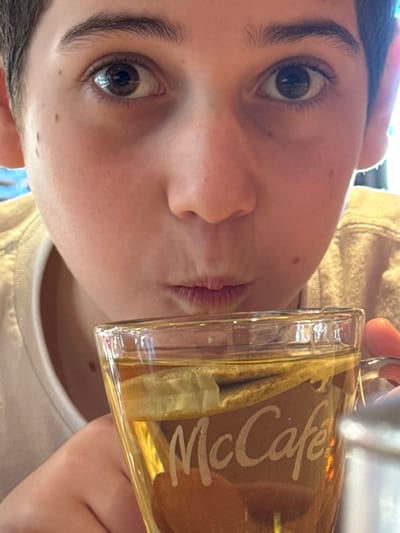 A close-up, eye-level image captures a young individual with fair skin, dark hair, and brown eyes, looking directly into the camera. Their mouth is puckered, suggesting either blowing on a beverage or making a specific facial expression. The individual is holding a clear glass mug with the "McCafé" logo etched on its side. The mug contains a light amber liquid and a submerged tea bag. A portion of the individual's right hand is visible on the mug handle. The subject is wearing a light-colored t-shirt, with a light brown garment partially visible on the right. The background is blurred, indicating an indoor setting. This scene is located in Lipomo, Italy.