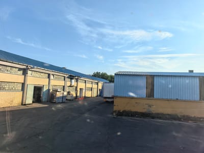 The image captures an exterior view of a warehouse facility under a bright, partly cloudy sky. In the foreground, a cracked asphalt lot is visible. To the left, a long, yellow building with a blue metal roof features numerous glass-block windows and at least two large, open bay doors. Pallets of goods are stacked outside one of these doors. A semi-trailer truck with a white trailer is parked to the right of this building, partially obscured by a second, blue-sided structure. This second structure also has a blue metal roof and a yellow base. The scene appears to be in an industrial area, possibly on the outskirts of the city of Osijek, Croatia, given the context provided. No people are clearly discernible in the image, though a figure may be present within the open bay door.
