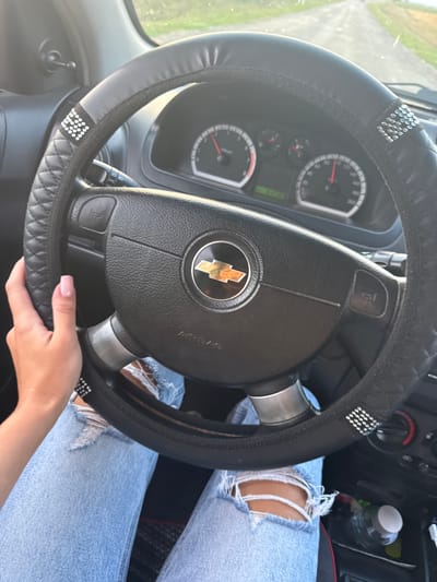 A close-up, first-person perspective shot captures the interior of a car, focusing on the steering wheel and dashboard. A person's hand with a light pink manicure is visible on the left side of the steering wheel, which is covered with a black, quilted material adorned with rhinestones. The car's dashboard includes a speedometer and tachometer, with the needles indicating movement. The driver is wearing distressed light-wash denim jeans. A partially visible water bottle rests in a cup holder. The road ahead is visible through the windshield, suggesting the car is in motion. The scene appears to be a country road, possibly in or near Sadaclia, Moldova, given the context provided.