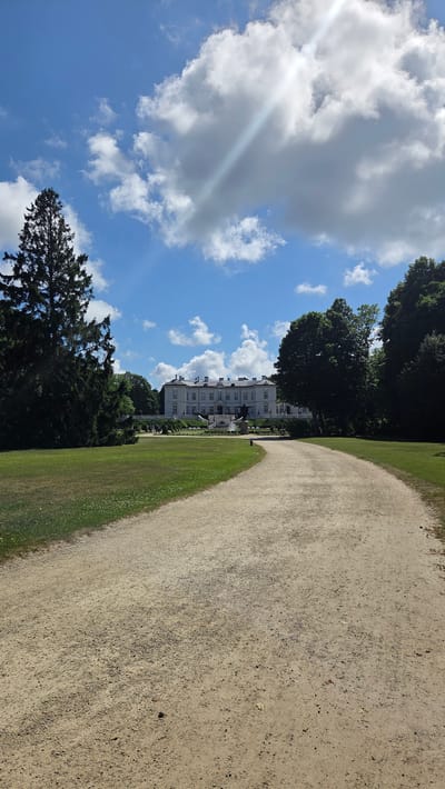 An eye-level, vertical, medium shot captures a sunlit path leading towards the Raudonvaris Manor, also known as the Tiškevičiai Palace, located in Palanga, Lithuania. The manor, a grand, white, neoclassical building with multiple wings and a prominent central section, sits atop a gentle rise, flanked by lush green lawns and dense trees. A wide, sandy path dominates the foreground, curving to the right and appearing to lead directly to the manor's grand staircase. The sky is a vibrant blue, dotted with fluffy white clouds, with a bright sun casting diagonal rays across the scene. The grounds are manicured, with well-kept grass and scattered trees, including a tall, dark green conifer on the left side of the frame. Several figures are visible on the grounds, scattered between the path and the manor, suggesting it is a public space. The overall atmosphere is serene and picturesque, typical of a well-maintained estate on a clear day.