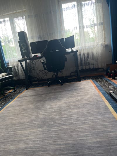 The image shows a room with a desk, chairs, and multiple computer monitors positioned in front of a window with sheer curtains. On the desk, there is a computer tower and other electronic equipment. To the left of the desk, there is a green gaming chair with a white cat sleeping on it. To the right of the desk, there are dumbbells and part of a treadmill. The room appears to be a home office or gaming setup. The location is identified as Aqköl, Kazakhstan.