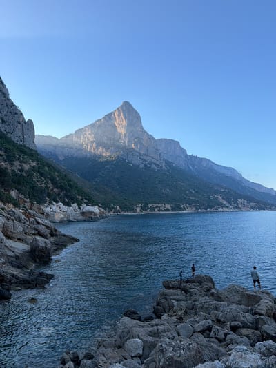 The image depicts a coastal landscape with rugged, rocky terrain meeting a calm, blue sea. Towering, sun-drenched mountains form the backdrop, with dense green vegetation clinging to their lower slopes. In the foreground, large, grey rocks are scattered along the shoreline and jut into the water. Three individuals are visible standing on a cluster of these rocks. One person, wearing a light-colored shirt and dark shorts, stands facing away from the camera, looking out at the water. Two other individuals, dressed in darker clothing, are positioned slightly behind and to the left of the first person. The sky is clear and a bright, pale blue. The scene evokes a sense of natural beauty and tranquility, likely in a Mediterranean setting. While the prompt requests a specific location in Baunei, Italy, this information cannot be definitively determined from the visual elements alone.